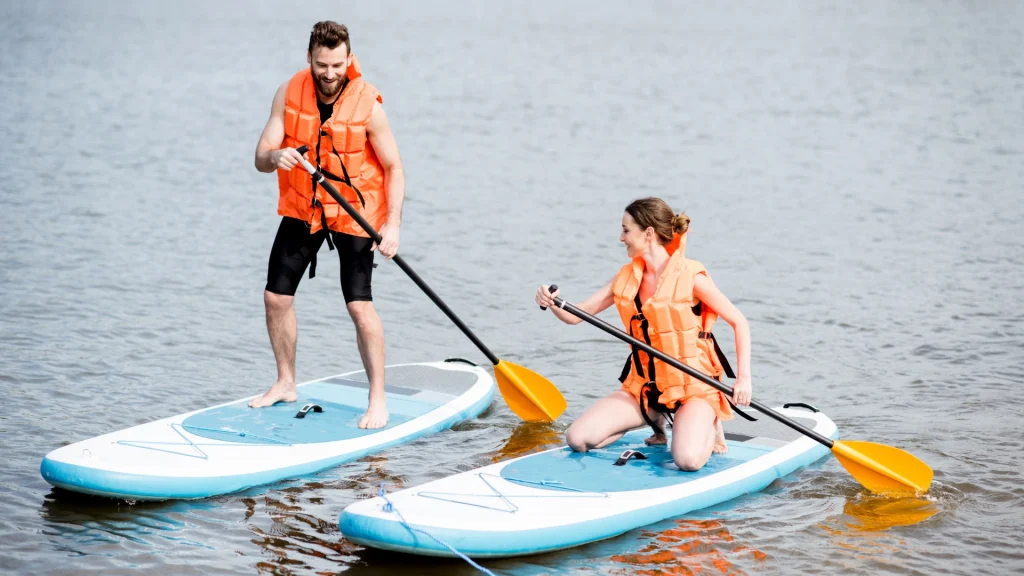 Két narancssárga mentőmellényt viselő ember élvezi a SUP evezést egy nyugodt tavon. A férfi mosolyogva áll, miközben egy fekete lapátot markol, míg a nő ülve nevet, és egy hasonló fekete lapáttal evez. Mindketten a part közelében vannak, és élvezik ezt az álló evezős kalandot.