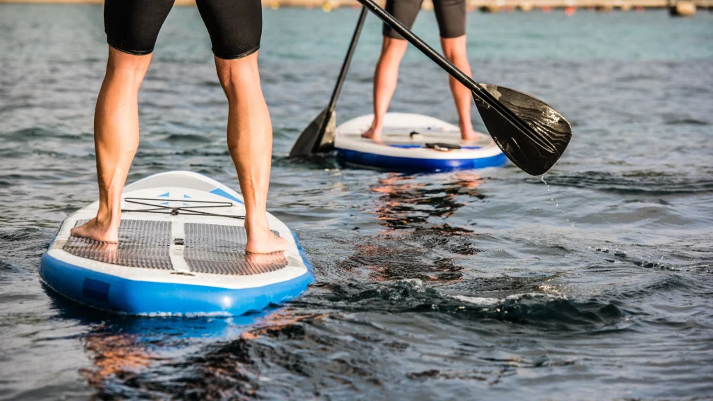 Két ember SUP evezést élvez egy nyugodt vízparton. Kék-fehér táblákon állnak, fekete rövidnadrágot viselnek, alsó testük látható. A víz finoman hullámzik a tiszta égbolt alatt, és egy távoli partvonal keretezi a derűs jelenetet.