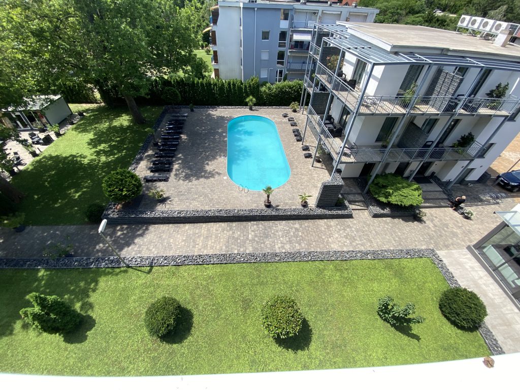 A felülről lefelé néző kilátás egy téglalap alakú medencére, amelyet nyugágyakkal ellátott terasz vesz körül, és nyugodt menekülést kínál. A medence mellett található egy modern, háromszintes, erkélyes épület, amely kiválóan alkalmas a SUP evezés szerelmeseinek megfigyelésére. A tájat szépen nyírt fű és apró cserjék jellemzik, miközben egy ember sétál a közelben.