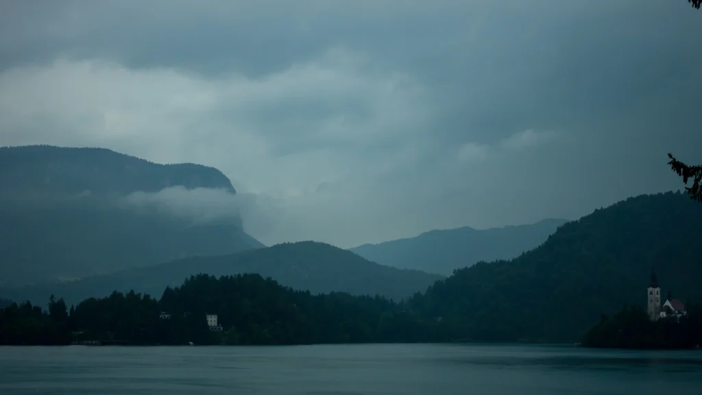 A nyugodt táj előtérben egy nyugodt tó található, amelyet ködös, erdős dombok vesznek körül a borús égbolt alatt. A part melletti kis kápolna sziluettje időtlen varázst tükröz, ahogy a hegyek rétegei visszahúzódnak a távolba. Ez a festői jelenet elmélkedésre és Időjárás Ellenőrzése hív.