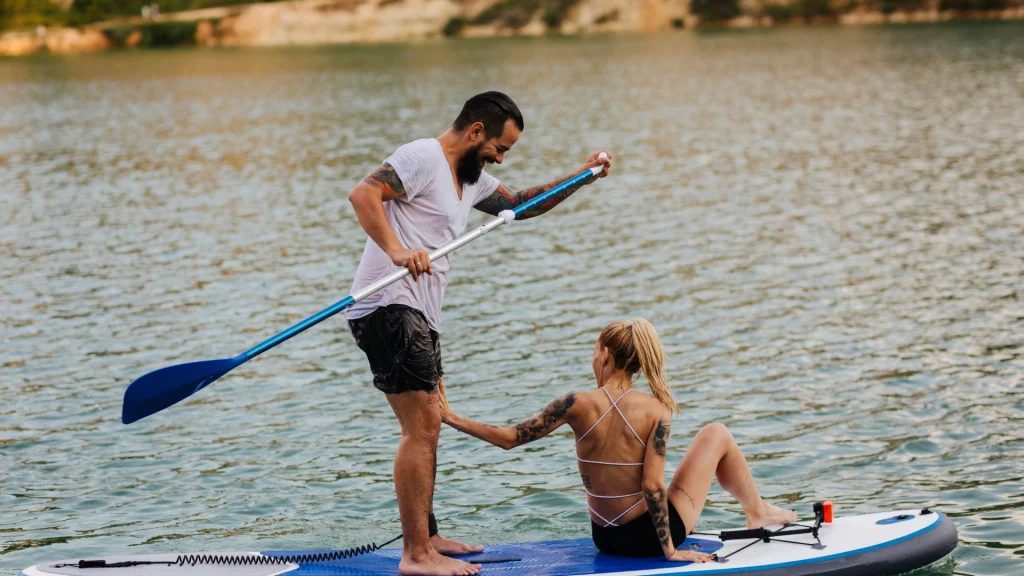 Egy szakállas férfi magabiztosan áll az evezős deszkán, megtestesítve a SUP biztonságot, miközben felsegít egy ülő nőt. Mindketten tetoválásokat mutatnak be, miközben nyugodt víz és buja növényzet veszi körül őket egy sziklás parton. Egy lapát a kezében, fürdőruhájában ellazul, biztonságban.