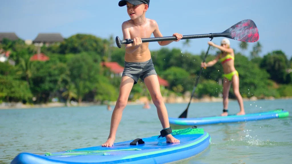 Egy fiatal, sapkás és rövidnadrágos fiú nyugodt vízen evez, oldalt tartva az evezőt. Mezítláb, bokasíjjal élvezi az egyik legjobb deszkát, miközben a háttérben egy másik bikinis evez. Zöld fák és épületek állnak a tiszta kék ég alatt. SUP Deszkák