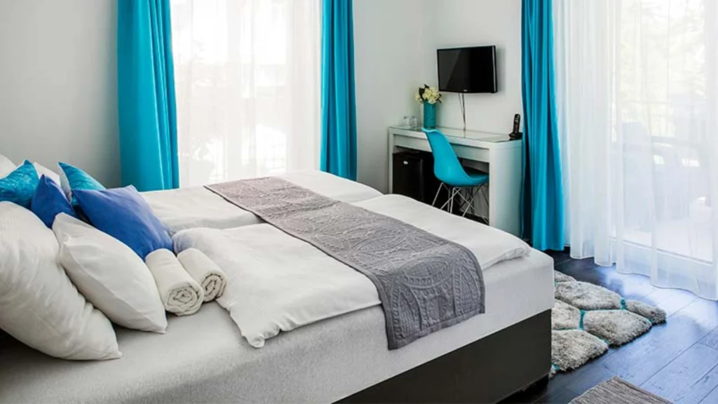 Világos, modern hálószoba szépen megvetett ággyal, fehér ágyneművel, kék-fehér párnákkal és szürke futószalaggal. A törölközők az ágy lábánál vannak feltekerve. A szoba elrendezése a stílus és a funkcionalitás automatikus vázlatát kínálja.