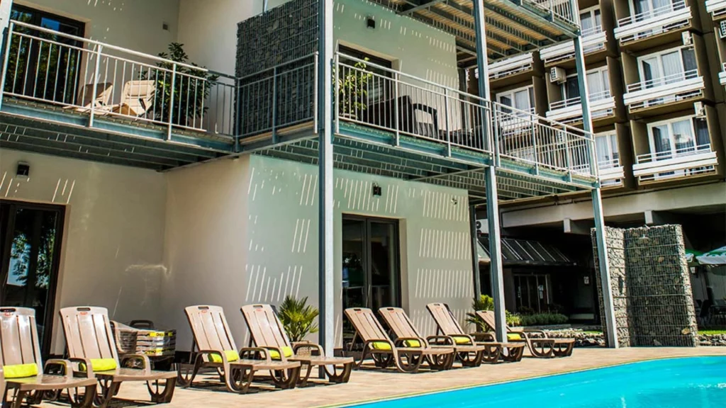 Külső medenceparti rész egy modern szállodában, ahol több barnás, sárga párnás nyugágy sorakozik a fedélzeten, szinte automatikus vázlatnak tűnő elrendezésben. Mögötte erkélyes szobák, fém korlátok, kő és fehér külső falak találhatók.