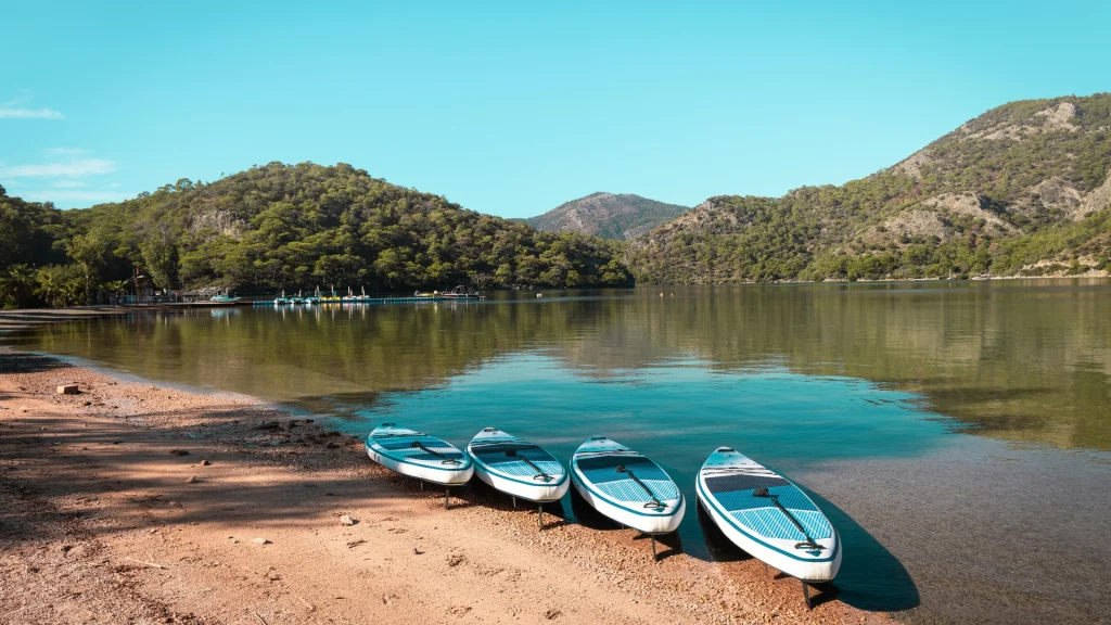 Nyugodt tóparti jelenetben négy SUP deszkák típusai pihennek egy homokos tengerparton. A tiszta víz tükrözi a buja zöld dombokat és a tiszta kék eget. Magas, erdős hegyek magasodnak a távolban, békés és festői szabadtéri környezetet teremtve.
